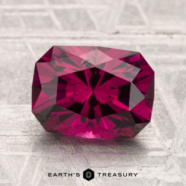3.10-Carat Umbalite Garnet A 3.10-carat Umbalite Garnet in deep pink rests on a textured gray surface, with "Earth's Treasury" and a hexagon logo at the bottom.