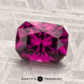A 2.54-carat Umbalite garnet, featuring a faceted deep pink hue, is displayed on a textured grey surface with the text "EARTH'S TREASURY" below it.