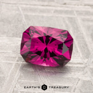 A 1.79-carat Umbalite garnet with a faceted, deep pink hue rests on a textured light gray surface, with "EARTH'S TREASURY" at the bottom.