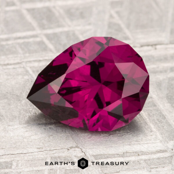 2.72-Carat Umbalite Garnet A 2.72-carat Umbalite Garnet, faceted in a pear shape and displaying a vivid magenta hue, rests on a textured gray surface stamped with the Earth's Treasury logo.