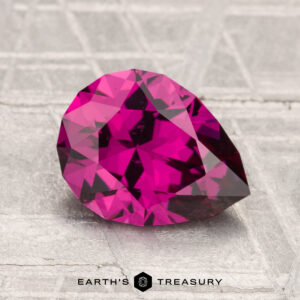 A faceted 2.13-carat Umbalite Garnet in deep pink rests on a textured metallic surface with "Earth’s Treasury" text below.