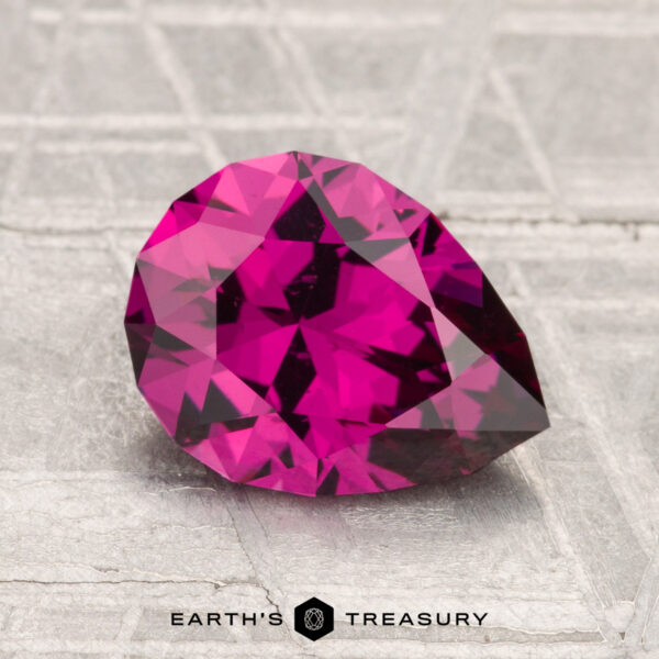 2.13-Carat Umbalite Garnet A faceted 2.13-carat Umbalite Garnet in deep pink rests on a textured metallic surface with "Earth’s Treasury" text below.