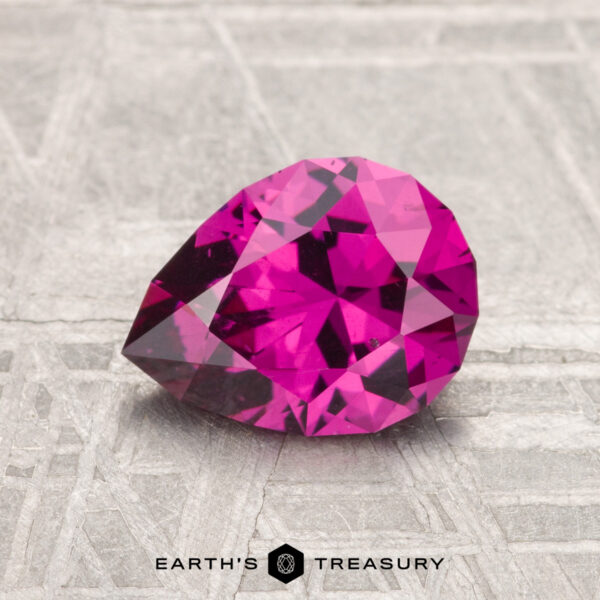 1.57-Carat Umbalite Garnet A faceted, pear-shaped 1.57-carat Umbalite garnet with a deep pink hue rests on a textured gray surface, with the words "EARTH'S TREASURY" beneath.