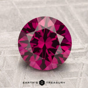 A 2.82-carat Umbalite Garnet, round and faceted in deep pink, rests on a textured gray surface with "EARTH'S TREASURY" at the bottom.