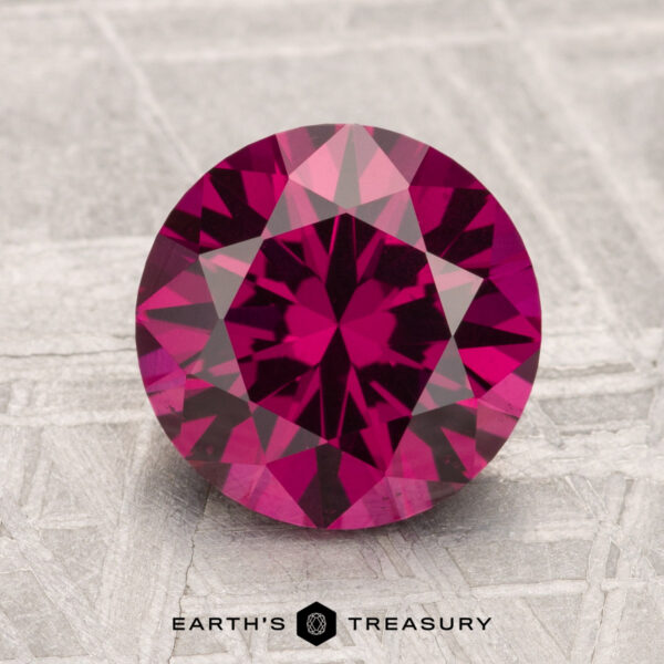 2.82-Carat Umbalite Garnet A 2.82-carat Umbalite Garnet, round and faceted in deep pink, rests on a textured gray surface with "EARTH'S TREASURY" at the bottom.