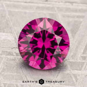 A 2.56-carat faceted Umbalite Garnet in deep pink is displayed on a textured gray surface with "Earth's Treasury" text below.