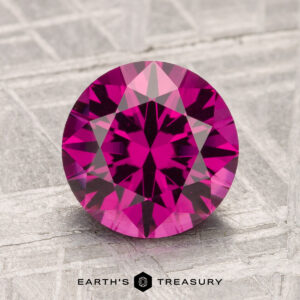 A 2.42-carat Umbalite Garnet with a round brilliant cut is displayed on a textured gray surface, with the "EARTH'S TREASURY" text and logo beneath it.