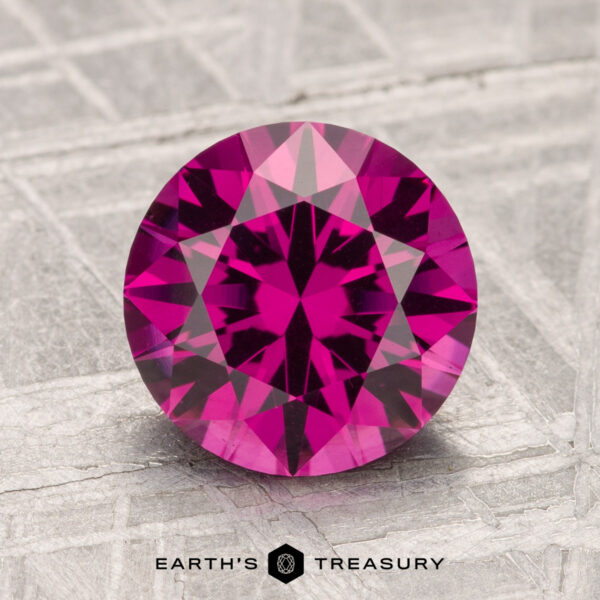 2.42-Carat Umbalite Garnet A 2.42-carat Umbalite Garnet with a round brilliant cut is displayed on a textured gray surface, with the "EARTH'S TREASURY" text and logo beneath it.