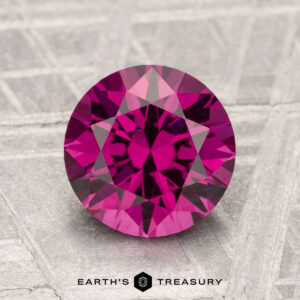 A 2.20-carat round, faceted deep pink Umbalite Garnet is displayed on a textured light gray surface with the text "Earth's Treasury" below.