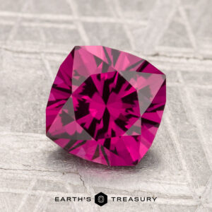 A 1.99-carat Umbalite Garnet, cushion-cut with vivid pink hues and geometric facets, rests on a textured metallic surface. "EARTH'S TREASURY" appears at the bottom of the image.