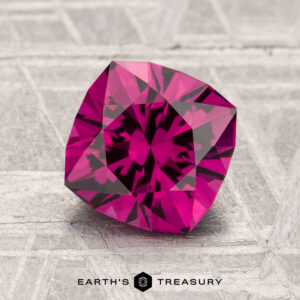 A 1.95-carat cushion-cut Umbalite Garnet with intricate facets rests on a textured gray surface, with "Earth's Treasury" displayed below.
