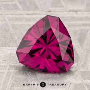 A 2.64-carat Umbalite Garnet, faceted in a triangular shape and featuring a deep pink-red hue, rests on a textured gray surface with the "Earth's Treasury" logo at the bottom.