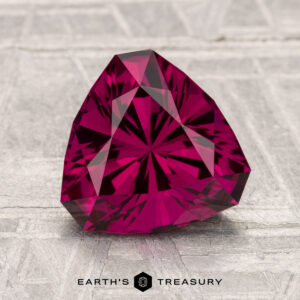 A 2.64-carat Umbalite Garnet in a deep pink trillion cut rests on a textured, light surface with "Earth's Treasury" beneath it.