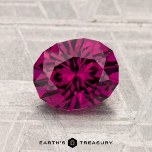 A faceted, oval-cut 1.70-carat Umbalite Garnet with a deep pink hue is displayed on a textured gray surface, with "Earth's Treasury" written below.