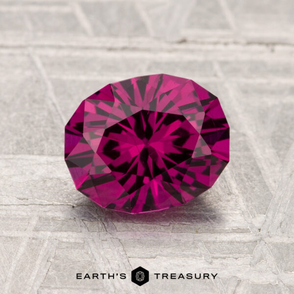 A faceted, oval-cut 1.70-carat Umbalite Garnet with a deep pink hue is displayed on a textured gray surface, with "Earth's Treasury" written below.