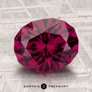A 3.70-carat Umbalite Garnet with symmetrical cuts and deep red hues rests on a textured surface, reflecting light. The text "EARTH’S TREASURY" appears at the bottom.