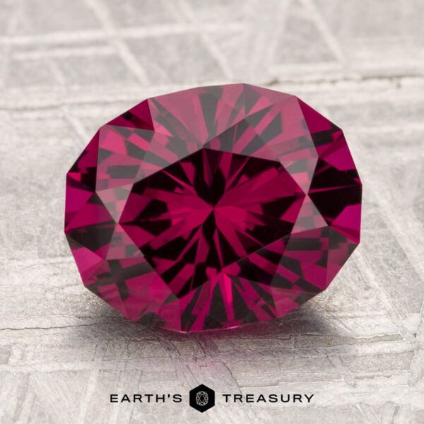 3.70-Carat Umbalite Garnet A 3.70-carat Umbalite Garnet with symmetrical cuts and deep red hues rests on a textured surface, reflecting light. The text "EARTH’S TREASURY" appears at the bottom.