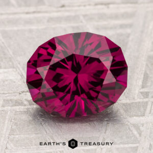 A round, faceted 3.16-carat Umbalite Garnet with a deep pink-red hue is shown on a textured, light background, with "EARTH'S TREASURY" written beneath.