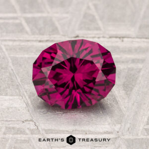 A 1.61-carat oval-cut Umbalite Garnet with a deep pink hue rests on a textured silvery-gray surface, featuring the text "Earth's Treasury" at the bottom.