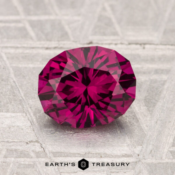 1.61-Carat Umbalite Garnet A 1.61-carat oval-cut Umbalite Garnet with a deep pink hue rests on a textured silvery-gray surface, featuring the text "Earth's Treasury" at the bottom.