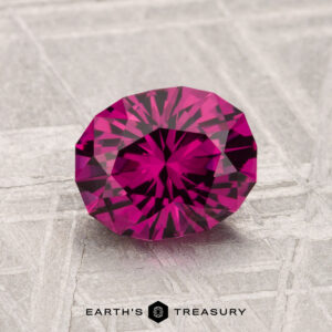 A 1.61-carat Umbalite garnet with a round cut and intricate facets is displayed on a textured gray surface near the Earth’s Treasury logo.