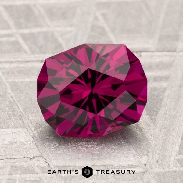 2.38-Carat Umbalite Garnet A 2.38-carat Umbalite Garnet, deep red and faceted, rests on a textured metallic surface with "EARTH'S TREASURY" displayed below.