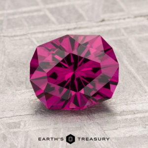 A 2.20-carat Umbalite Garnet with a geometric faceted cut in deep pink rests on a textured gray surface, with "Earth's Treasury" shown at the bottom of the image.