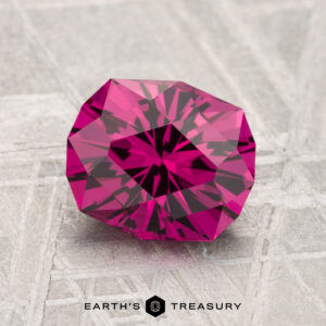 A 1.95-carat Umbalite Garnet, round brilliant-cut and deep pink, is displayed on a textured gray surface with the "Earth's Treasury" logo beneath it.