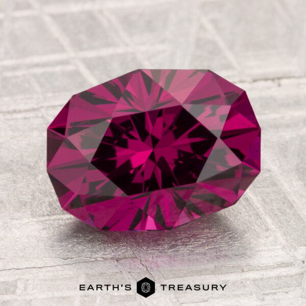 3.60-Carat Umbalite Garnet A 3.60-carat Umbalite Garnet, faceted in an oval cut with a deep pink hue, rests on a light textured surface with "EARTH'S TREASURY" text displayed at the bottom.
