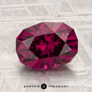 A 3.13-carat Umbalite Garnet, faceted in deep red, rests on a textured light surface with the "Earth's Treasury" logo and text below it.