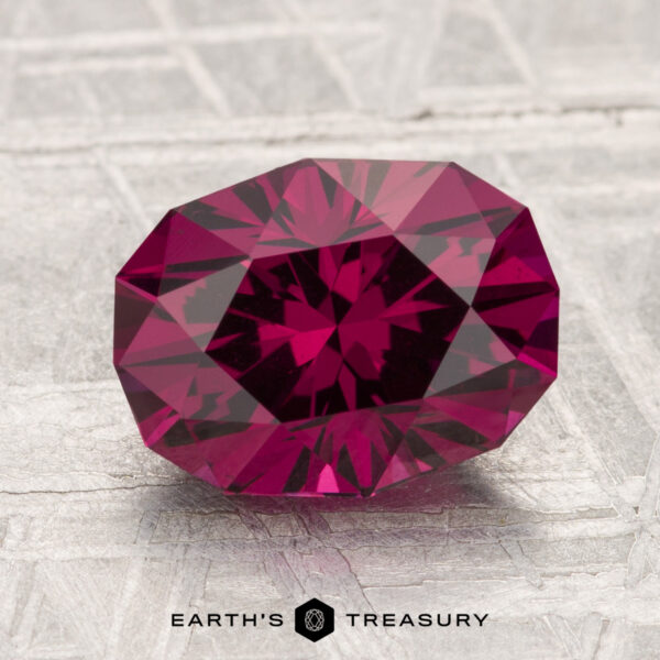 3.13-Carat Umbalite Garnet A 3.13-carat Umbalite Garnet, faceted in deep red, rests on a textured light surface with the "Earth's Treasury" logo and text below it.