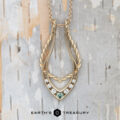 The "April" ring keeper necklace in 14K yellow gold, brushed finish, features twisted and smooth textures with five clear stones and one green stone. Presented on a light textured background with "Earth's Treasury" text below.