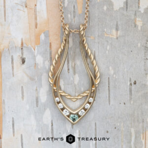 The "April" ring keeper necklace in 14K yellow gold, brushed finish, features twisted and smooth textures with five clear stones and one green stone. Presented on a light textured background with "Earth's Treasury" text below.