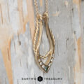 The "April" ring keeper necklace in brushed 14K yellow gold is displayed against birch bark, featuring a twisted design with small diamonds and a green gemstone. The "Earth's Treasury" logo appears at the bottom.