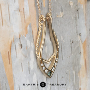 The "April" ring keeper necklace in brushed 14K yellow gold is displayed against birch bark, featuring a twisted design with small diamonds and a green gemstone. The "Earth's Treasury" logo appears at the bottom.