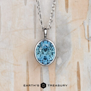 The "Foggy" pendant in 14k white gold features a 1.37-carat oval Montana sapphire on a chain, shown against a light, textured background. "EARTH'S TREASURY" logo and text are at the bottom.