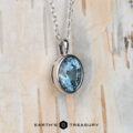 The "Foggy" pendant in 14k white gold features a 1.37-carat oval Montana sapphire, elegantly displayed against a light wood background with the Earth’s Treasury logo at the bottom.