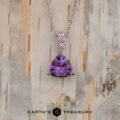 The "Noelani" necklace in 14K white gold features a 4.03-carat triangular amethyst and a small round pink gemstone above it, displayed against a light, textured wood background.