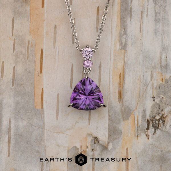 The "Noelani" necklace in 14K white gold with 4.03-Carat amethyst The "Noelani" necklace in 14K white gold features a 4.03-carat triangular amethyst and a small round pink gemstone above it, displayed against a light, textured wood background.