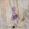 The "Noelani" necklace in 14K white gold showcases a 4.03-carat triangular amethyst and two small round purple stones, displayed on light wood with "EARTH'S TREASURY" at the bottom.