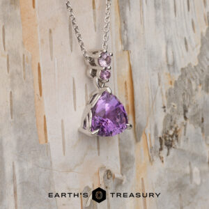 The "Noelani" necklace in 14K white gold showcases a 4.03-carat triangular amethyst and two small round purple stones, displayed on light wood with "EARTH'S TREASURY" at the bottom.