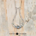 The "Twilight" ring keeper necklace in 14K white gold features a teardrop-shaped pendant with six blue and green gemstones, displayed on light wood with “EARTH’S TREASURY” text below.