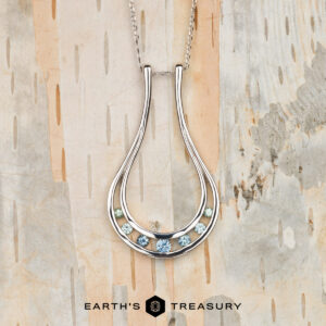 The "Twilight" ring keeper necklace in 14K white gold features a teardrop-shaped pendant with six blue and green gemstones, displayed on light wood with “EARTH’S TREASURY” text below.