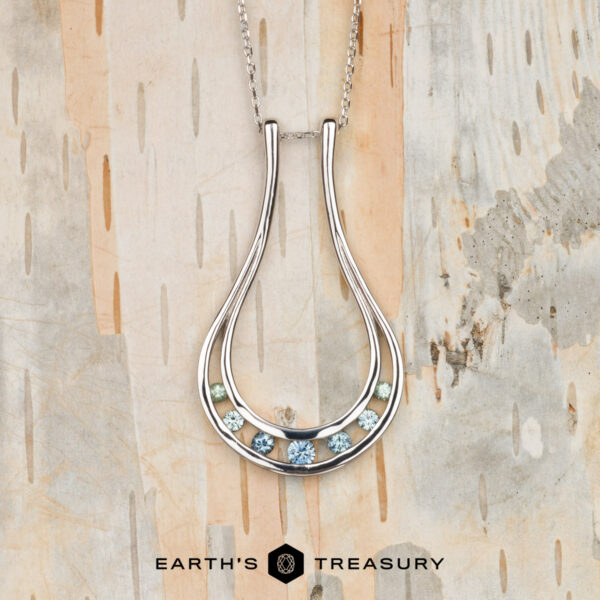 The "Twilight" ring keeper necklace in 14K white gold features a teardrop-shaped pendant with six blue and green gemstones, displayed on light wood with “EARTH’S TREASURY” text below.