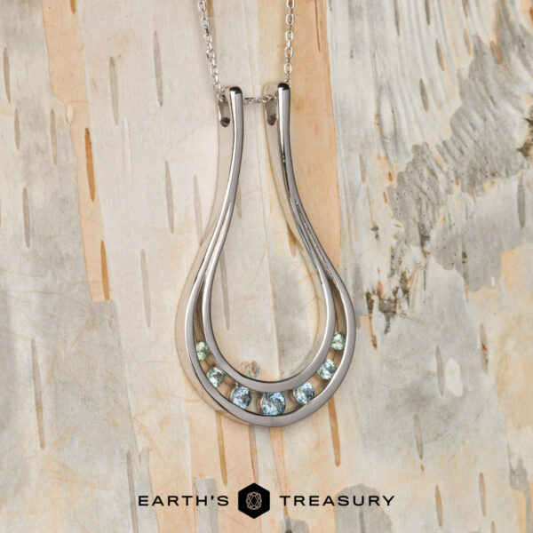 The "Twilight" ring keeper necklace in 14K white gold The "Twilight" ring keeper necklace in 14K white gold features a teardrop design with seven small gemstones at the base, shown on a light wooden background with the "Earth's Treasury" logo below.