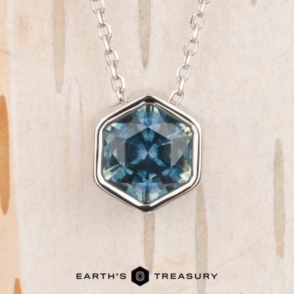 The Build Your Own "Shimmer" pendant in 14k white gold with 1.62-carat Montana sapphire The Build Your Own "Shimmer" pendant in 14k white gold features a 1.62-carat Montana sapphire, shown against a light textured background with the "Earth's Treasury" logo at the bottom.