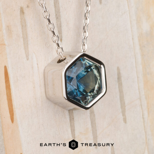 The Build Your Own "Shimmer" pendant in 14k white gold with 1.62-carat Montana sapphire The Build Your Own "Shimmer" pendant in 14k white gold features a 1.62-carat Montana sapphire, shown on a white gold chain against a light wooden background with the "Earth's Treasury" logo at the bottom.