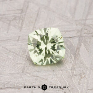 A 0.59-Carat Grossular Garnet with a cushion cut and pale mint green hue is shown on a textured gray surface, featuring the "EARTH'S TREASURY" logo and text at the bottom.