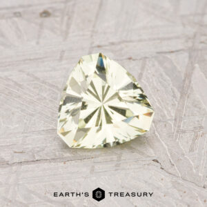 A pale mint green, trillion-cut grossular garnet weighing 0.83 carats rests on a textured light gray surface with the text "Earth's Treasury" at the bottom.