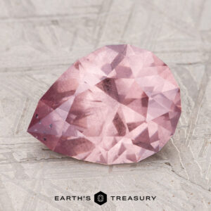 A 3.12-carat pink, pear-shaped faceted garnet rests on a textured surface with "Earth's Treasury" at the bottom.